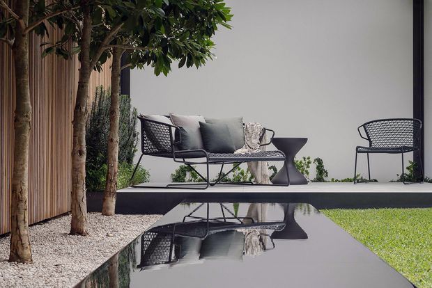 A refined outdoor terrace beside a modern residence, featuring a black outdoor lounge chair and sofa with cushions, a sculptural side table, and a matching chair arranged on a raised platform. Small trees and white gravel border the seating area, while a glossy dark reflecting pool or water feature mirrors the furniture. The setting has a restrained, contemporary palette of charcoal, grey, timber brown, white gravel, and green planting.