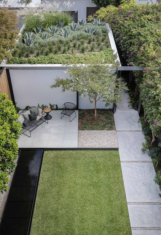 A contemporary landscaped courtyard viewed from above, featuring a lush planted rooftop or raised garden bed, sculptural succulents and ornamental grasses, a paved outdoor seating terrace with black wire furniture, a narrow dark reflecting pool, manicured lawn, and large-format concrete stepping slabs. White rendered walls and dense greenery create a refined urban garden setting.