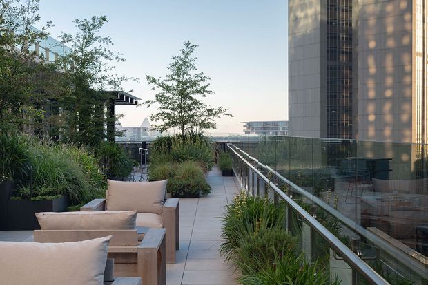 A landscaped rooftop terrace or elevated outdoor walkway beside a modern multi-storey building. The space features pale tiled paving, glass balustrades with metal handrails, large planters filled with ornamental grasses and trees, and built-in or substantial outdoor lounge seating with light timber frames and neutral cushions. Urban buildings are visible beyond the terrace, while the building facade at right has a warm-toned vertical panelled appearance.