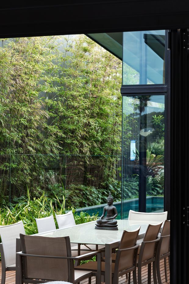 A refined indoor dining area beside full-height glazing, opening visually onto a lush tropical garden and narrow swimming pool. A large rectangular dining table with contemporary brown-framed chairs occupies a timber-floored space, with a Buddha statue as a table centrepiece. Dark window and door frames, extensive glass, and dense planting create a calm indoor-outdoor connection.
