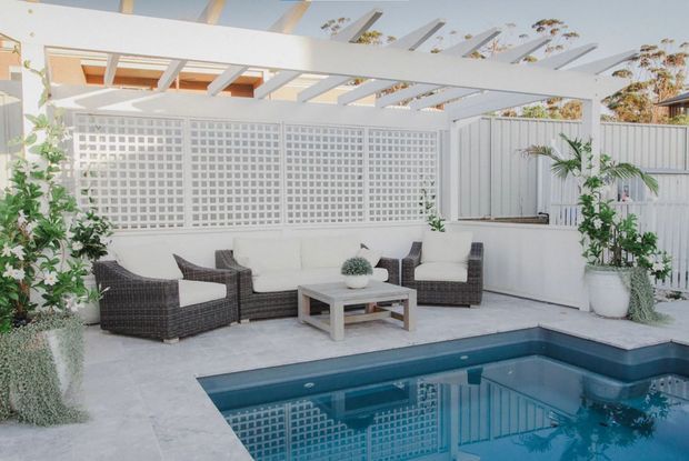 Bright, white-toned outdoor pool courtyard with a covered pergola, breeze-block privacy wall, tiled paving, and a casual wicker seating area arranged around a coffee table. Potted tropical greenery softens the structured enclosure, while the pool occupies the foreground.