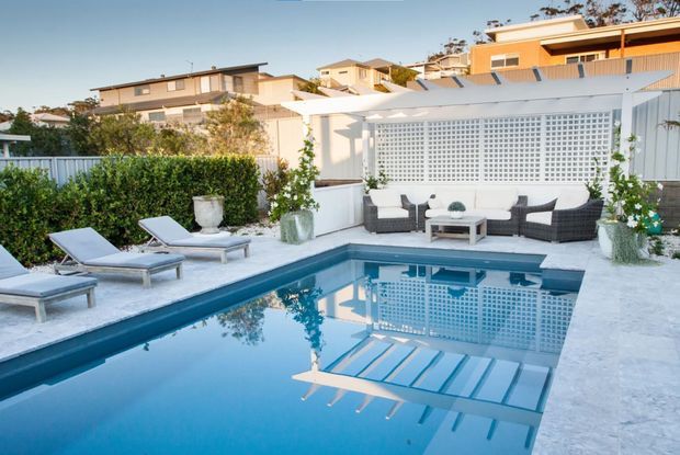 A bright outdoor residential pool area with a rectangular swimming pool, pale stone paving, sun loungers, and a black wicker seating arrangement. A white architectural screen wall with a breeze-block pattern forms the backdrop, with hedging, potted plants, and neighbouring homes visible beyond.