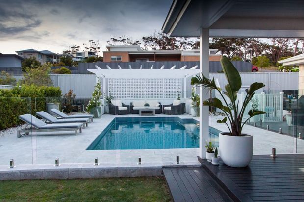 A landscaped residential backyard viewed from a covered deck, featuring a rectangular swimming pool, pale tiled surrounds, glass balustrades, sun loungers, and a white pergola with lattice screening and outdoor seating. Large tropical-style potted plants and neighbouring suburban homes are visible beyond the pool.