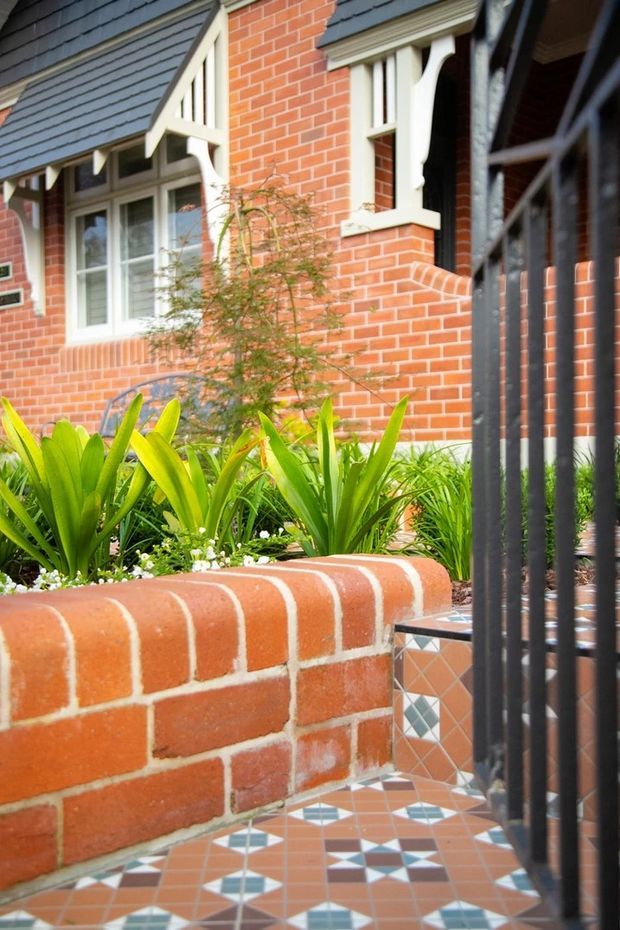 Close-up exterior view of a traditional red-brick house entrance, featuring white decorative timber window surrounds and gable detailing, dark slate-like roof cladding, a landscaped raised brick garden bed, black metal security gates, and a patterned terracotta-and-blue tiled entry path.