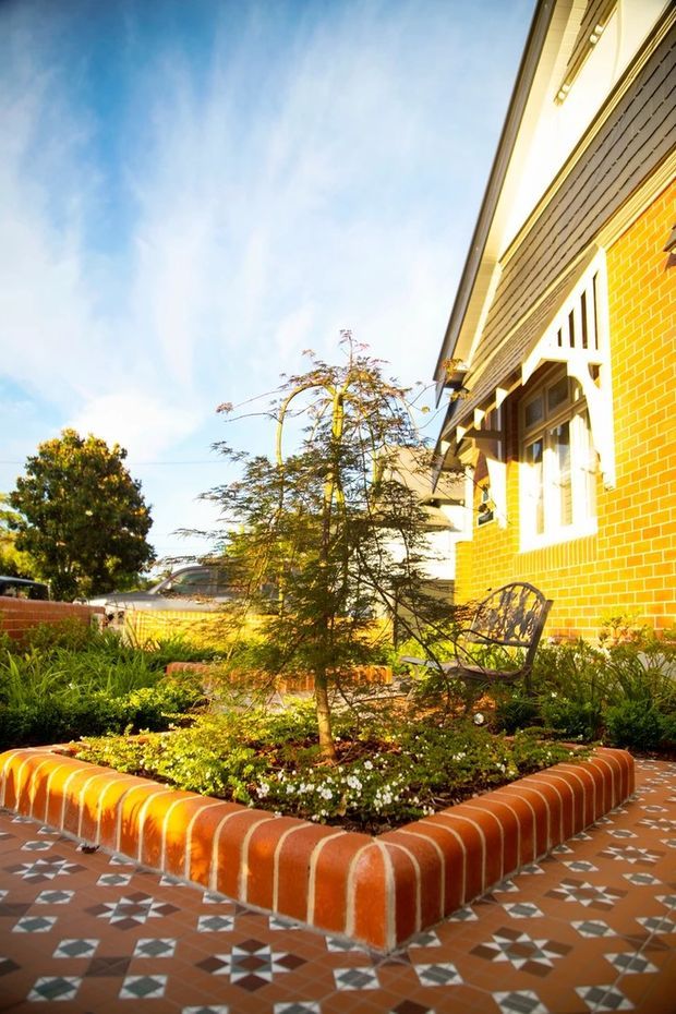 A sunlit residential courtyard garden beside a red brick house, featuring a raised brick planter with a small tree and low planting, patterned terracotta-and-white tiled paving, and a metal garden chair. The house has white-trimmed windows, decorative timber detailing, and a pitched roof edge.