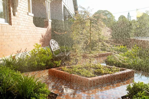 A sunlit landscaped courtyard beside a brick building, featuring a raised rectangular garden bed surrounded by shallow reflective water or wet paving, patterned terracotta-toned tiles, dense planting, and a white metal garden chair. The scene has a tranquil, established garden character with mature vegetation and masonry walls.