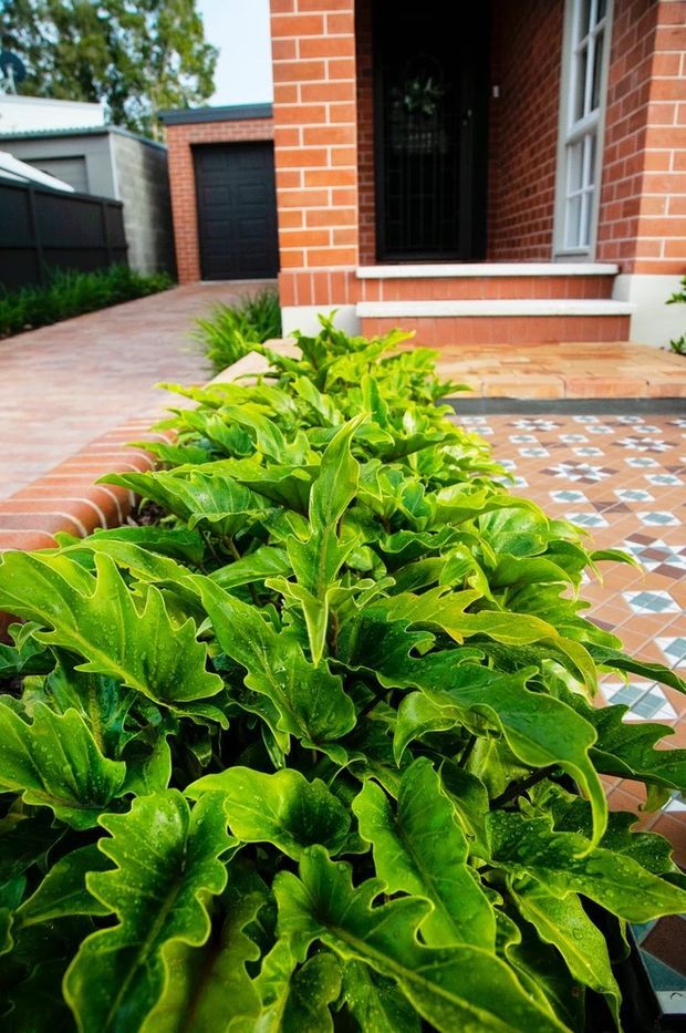 A residential front entry viewed from the garden path, featuring a red brick facade, black security door, white-trimmed windows, tiled entry steps and porch, a patterned geometric tile paving area, and lush green planting in the foreground. A detached garage and paved driveway are visible to the side.