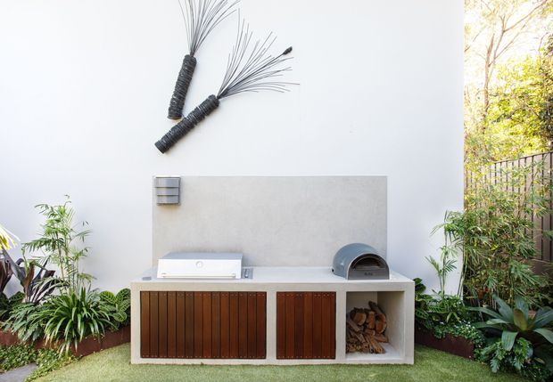 Outdoor entertaining area featuring a built-in barbecue and pizza oven set into a white rendered wall, with dark timber slatted cabinetry and an open firewood storage compartment. Lush planting surrounds the compact outdoor kitchen, while a black metal garden screen and trees are visible to the side. Decorative wire wall sculpture adds a contemporary touch.
