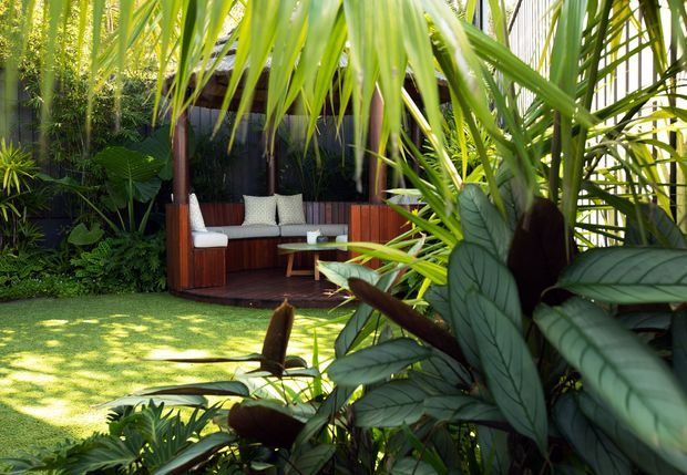 A lush tropical garden courtyard featuring a sheltered outdoor lounge pavilion with dark timber walls, vertical posts, built-in bench seating, and pale cushions. Dense broad-leaf planting surrounds a manicured lawn, creating a secluded resort-like outdoor retreat.