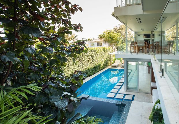 A bright contemporary multi-level residence viewed from a lush garden, with extensive floor-to-ceiling glazing, glass balustrades, white concrete surfaces, balconies, and a vivid blue swimming pool with stepping stones. Outdoor dining is visible on an upper terrace, while the lower level opens directly toward the pool.