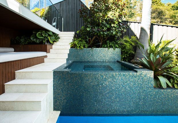 A lush landscaped outdoor courtyard featuring a compact tiled plunge pool or spa, with blue mosaic tile walls and basin, pale stone steps, dark timber-clad built-in seating, and dense tropical planting. The scene has a layered, contemporary indoor-outdoor design with strong geometric forms and a tranquil garden setting.