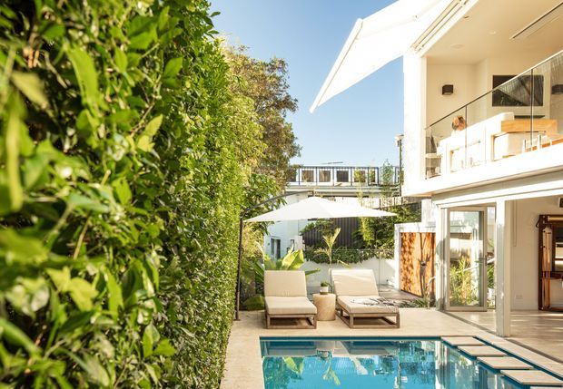 Sunny view of a contemporary multi-level home with a swimming pool, paved pool terrace, outdoor lounge furniture, large umbrella, glass balustrades, and lush screening hedges. The white facade features extensive glazing and projecting roof or awning forms, with interior spaces visible through the open glass doors.