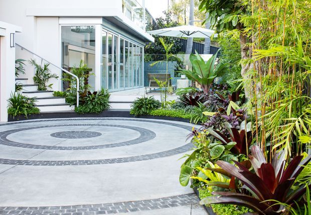 A contemporary residential courtyard or driveway with pale concrete paving and concentric dark stone inlay patterns. The space is bordered by dense tropical planting and leads to a white, glazed pavilion-like living area with broad windows, steps, and a large outdoor umbrella.