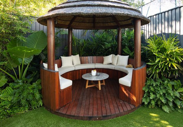 A lush landscaped outdoor lounge featuring a circular timber gazebo with a thatched roof, built-in curved upholstered seating, loose chairs, and a small round coffee table. Dense tropical planting surrounds the structure, with dark fencing forming the backdrop.