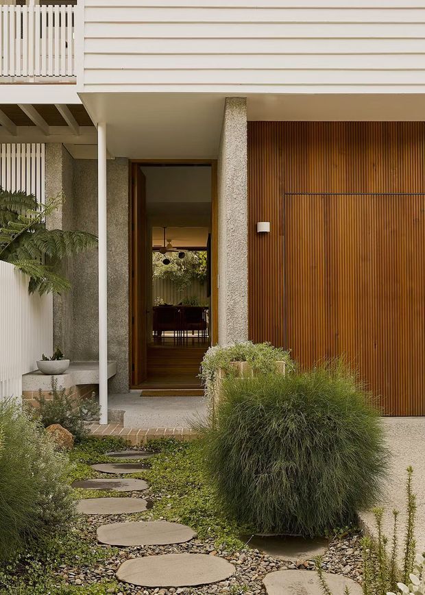 Front entry of a contemporary residential home, featuring a textured light render facade, horizontal cream cladding above, a warm timber slatted garage door, and a recessed timber-framed entry. A landscaped path of rounded concrete stepping stones runs through pebble groundcover and ornamental grasses, with a covered balcony and white vertical balustrade above.