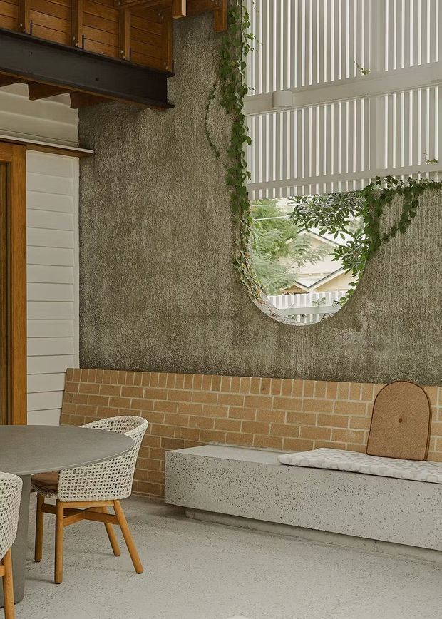 A distinctive covered outdoor dining and seating area with a round table, woven chairs, a long built-in concrete bench, exposed brickwork, rough textured plaster walls, timber detailing, climbing greenery, and a circular wall opening beneath a white vertical slatted screen. The space has a warm, tactile contemporary design with strong indoor-outdoor character.