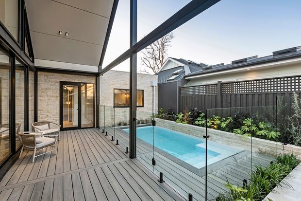 Contemporary covered outdoor deck and pool courtyard viewed from a glazed living area. The space features grey timber-look decking, slender dark-framed glass balustrades, a covered pergola-like roof structure, pale exposed brick walls, and a narrow rectangular swimming pool surrounded by landscaped planting and privacy screening.