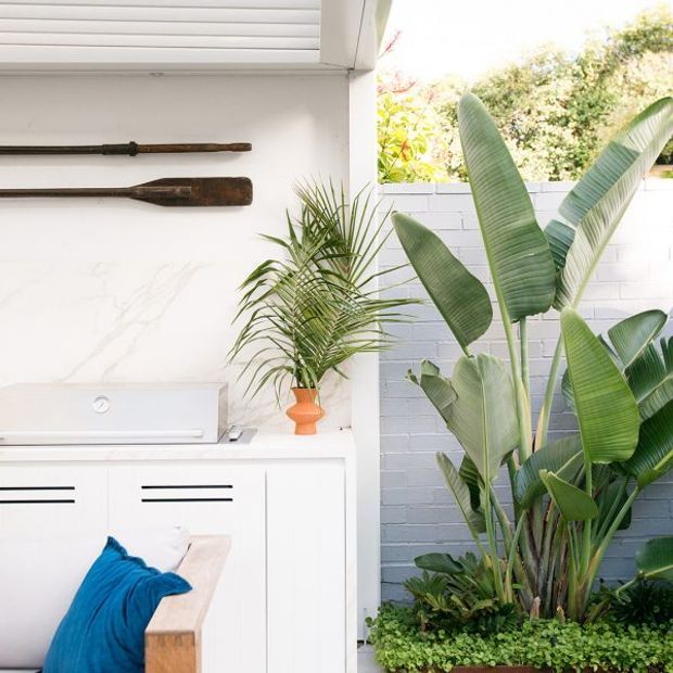 Bright, compact outdoor entertaining area beside a white built-in cabinet and barbecue, with a marble-look wall panel, decorative timber oars, and abundant tropical planting against a pale painted brick wall. A small bench with blue fabric adds a vivid accent.