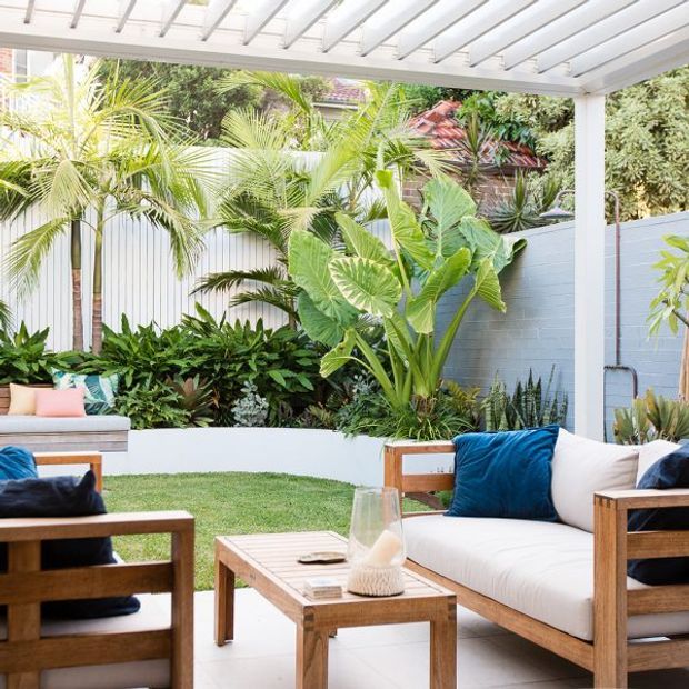 A bright outdoor lounge beneath a white slatted pergola, furnished with timber-framed sofas, white cushions, blue accent pillows, and a matching coffee table. The space opens onto a lush tropical courtyard garden with layered planting, palms, broad-leaf foliage, and a pale blue boundary wall.