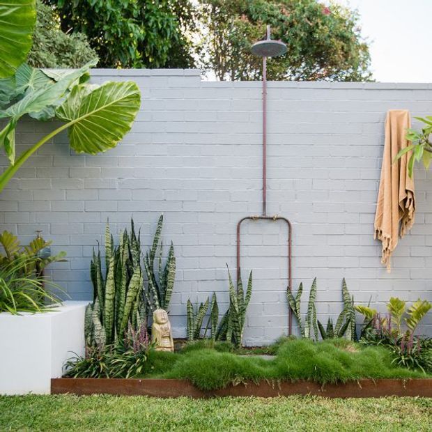 A lush landscaped outdoor shower area against a pale grey painted brick wall. A copper-toned exposed shower pipe and round showerhead are centered above a planted garden bed, with sculptural grasses, snake plants, broad tropical leaves, and a hanging tan towel. A white raised planter and lawn frame the foreground.