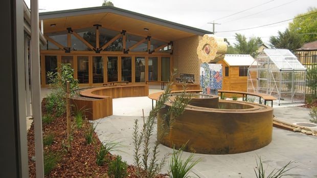 A landscaped courtyard or outdoor communal area with a timber-clad building, broad covered roof, extensive glazing, exposed structural bracing, curved timber seating, and large circular rusted metal planters or water features. A greenhouse and garden elements are visible to the right.