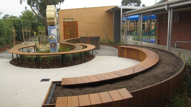 A landscaped courtyard or outdoor educational space with a winding curved timber bench and pathway surrounding planted garden beds. The setting includes exposed brick buildings, covered outdoor circulation, colourful sculptural or play elements, and glazed/open areas in the background. The design has a playful, organic layout with integrated seating and planting.