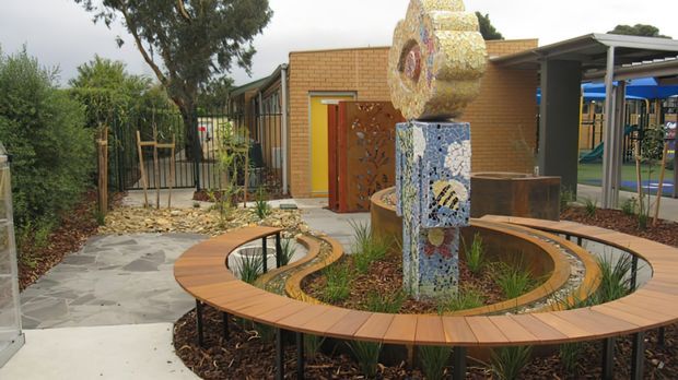A landscaped courtyard beside a low-rise brick building, featuring a circular timber bench surrounding a mosaic-tiled sculptural water or garden feature. Paved paths, planted beds, metal fencing, and a covered outdoor structure are visible.