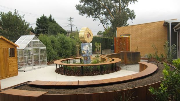 A landscaped courtyard with curved timber pathways and raised garden beds surrounding a circular planting area and sculptural feature. A small glass greenhouse and timber shed sit to the left, while a warm brick building forms the background. The space has a contemporary, organic garden design with flowing curves and integrated outdoor circulation.