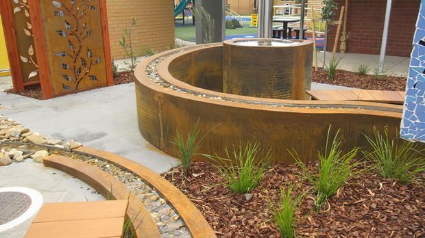 A landscaped outdoor courtyard featuring a curved corten-steel sculptural planter or raised garden bed, a matching circular central feature, timber decking, decorative metal screening, concrete paving, and ornamental grasses. The space has a contemporary public or commercial landscape-design character.