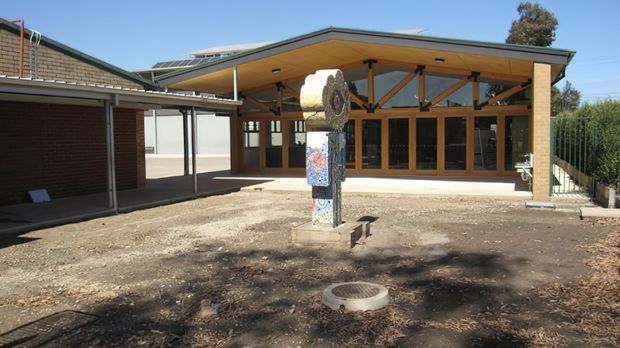 Single-storey timber-framed building viewed from an open courtyard, with a broad low-pitched roof, deep covered veranda, exposed timber trusses, extensive timber-framed glazing, and a central sculptural feature. An adjoining brick structure and covered link are visible to the left.