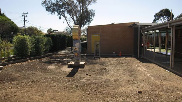 A sunny outdoor courtyard or school-like property area with a low red-brick building, covered walkway, fenced garden boundary, mature trees, and a central sculptural or utility column. The ground is mostly bare compacted earth, and the building has a simple flat or low-pitched roof with a yellow door.