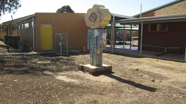 Exterior view of a low-rise institutional or school-like building surrounding a bare courtyard. The building has reddish-brown brick walls, covered walkways supported by slender metal posts, large glazed openings, and a bright yellow door. A colourful mosaic-like sculptural artwork stands in the centre foreground. The scene is sunlit with sparse ground cover and mature trees in the background.