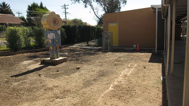 A sunlit outdoor courtyard or grounds area beside a low institutional-looking brick building. The unpaved, dusty surface surrounds a colourful decorative sculptural feature on a small concrete plinth, with a yellow service door, metal fencing, hedges, and covered structure along the right side. Residential rooftops and utility poles are visible beyond the fence.