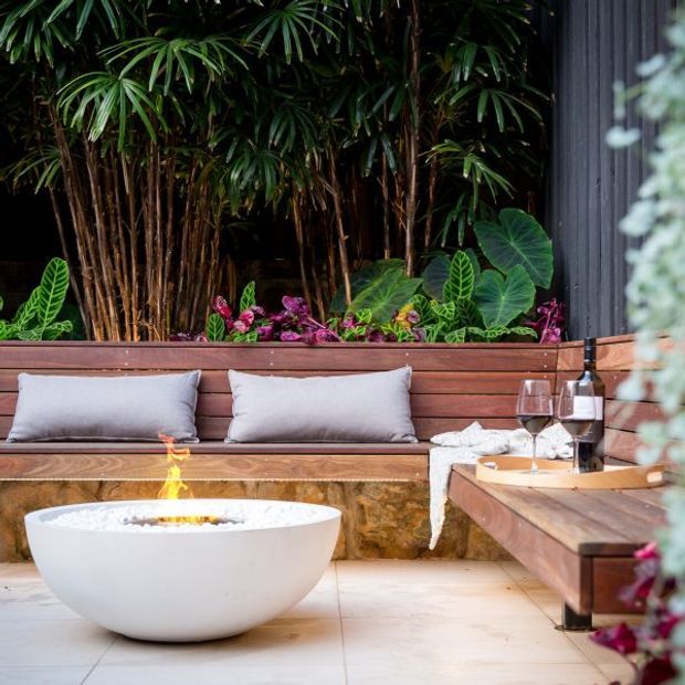 A lush outdoor lounge courtyard or terrace featuring a built-in timber bench with grey cushions, a white bowl-style fire pit, warm timber decking and joinery, dense tropical planting, and a small timber side table with wine glasses. The space has a refined, resort-like atmosphere with natural materials and strong greenery.