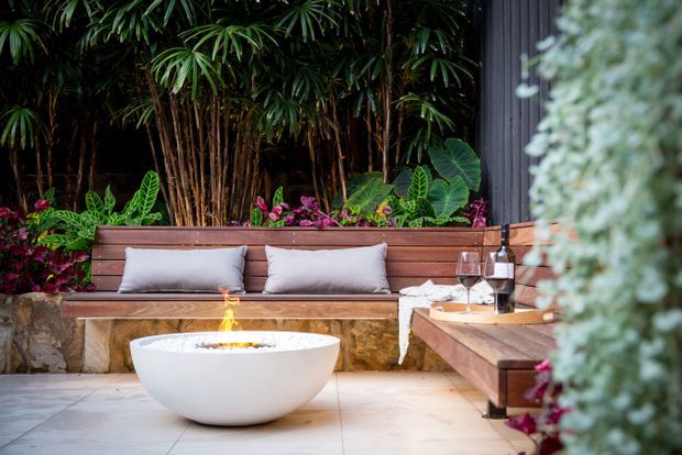 A lush, intimate outdoor lounge courtyard with a built-in timber bench lined with cushions, raised tropical planting beds, a low white fire bowl, and a timber-clad side bench or counter. The space has a warm, resort-like atmosphere with dark vertical cladding and pale tiled paving.