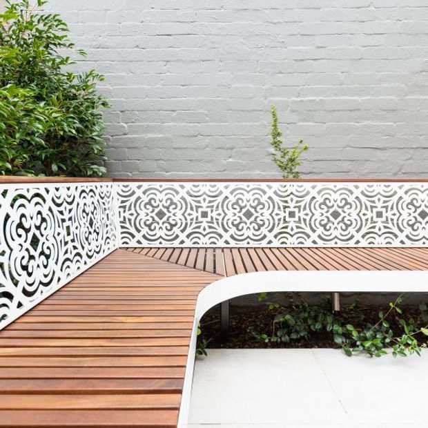 A compact landscaped courtyard or terrace featuring warm timber decking, ornate white perforated decorative screening along the edges, a curved white built-in bench or planter edge, a pale painted brick wall, and dense green planting. The image focuses on the hardscape and garden detailing.