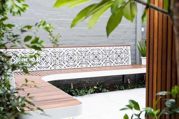 A leafy courtyard terrace with an L-shaped built-in timber bench surrounding a white paved area and planted garden bed. The bench backs onto a white-painted brick wall with an ornate white decorative screen, while timber vertical cladding frames the right side.