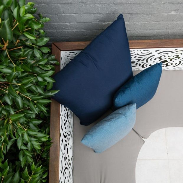 Top-down close-up of a styled outdoor or indoor lounge setting with navy and blue cushions arranged on a grey upholstered surface, framed by a dark timber border and ornate white patterned trim. Dense glossy green foliage occupies the left side, creating a layered, contrasting composition.