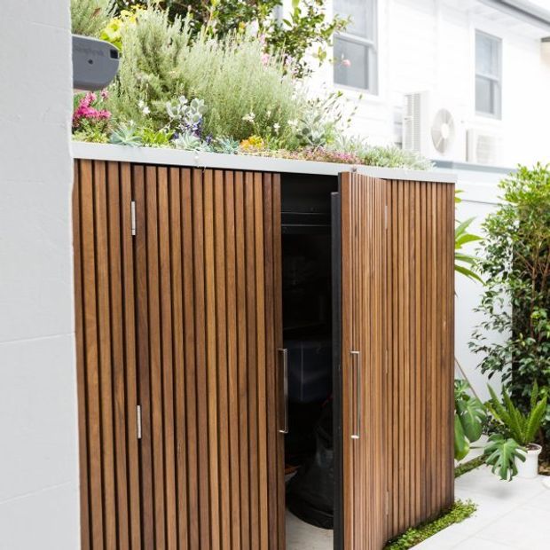 A compact outdoor storage cabinet with vertical timber slat doors and a planted green roof, positioned beside a white rendered building and landscaped courtyard. One door is open, revealing dark interior storage. The cabinet has a clean contemporary appearance and integrates greenery into its top surface.