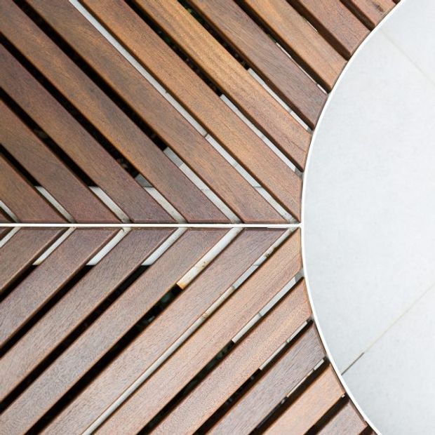 Close-up top-down view of a circular outdoor deck or platform made from radial timber slats, with dark gaps between the boards and a smooth pale curved surface forming the right edge. The composition emphasizes geometric radial patterning and natural wood grain.