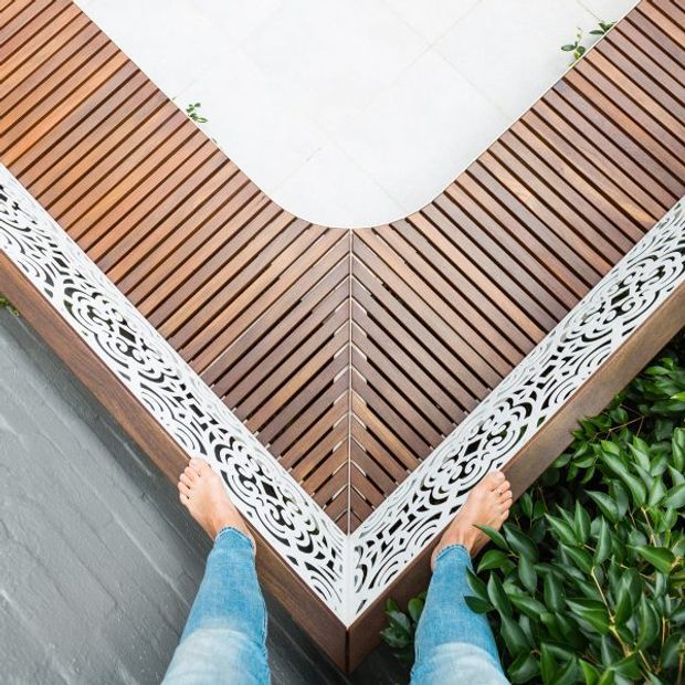 Top-down view of a small timber deck or balcony corner with diagonal slatted decking, a rounded white tiled inset, ornate white decorative privacy screens along the edges, and dense green planting beside a grey exterior wall. A person’s feet are visible, emphasizing the compact outdoor setting.