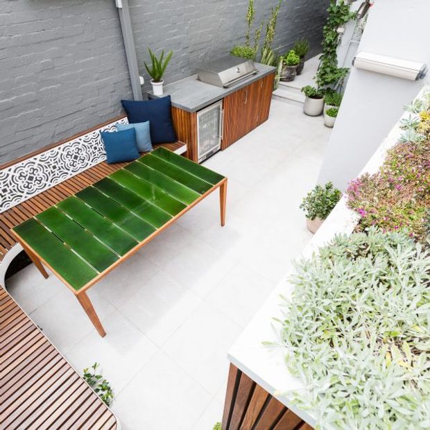 A compact urban courtyard or rooftop terrace with pale tiled flooring, grey painted brick walls, and abundant potted planting. The space includes a built-in outdoor kitchen with timber slatted cabinetry, stainless-steel benchtop and barbecue, a green outdoor dining table, and a timber bench with patterned metal backrest and blue cushions. A timber deck edge and raised planter filled with succulents complete the contemporary outdoor setting.