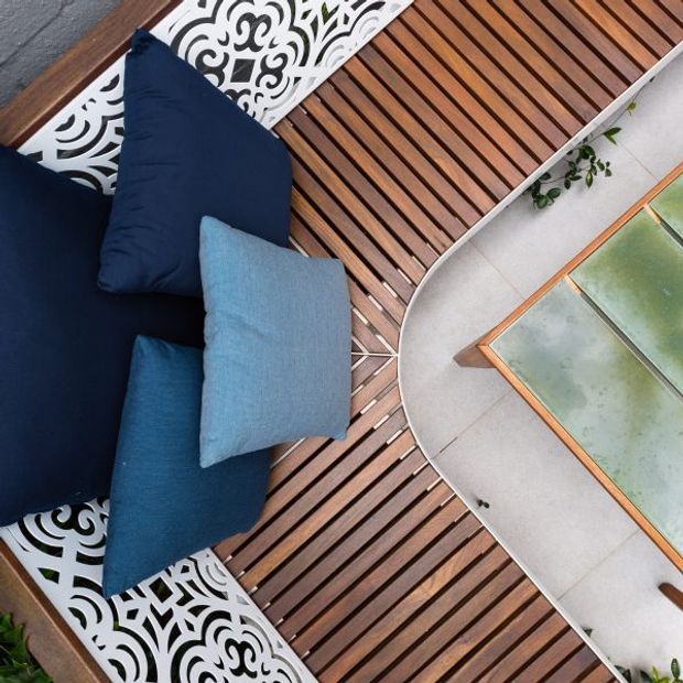Overhead view of a compact outdoor lounge area with deep navy and blue cushions on ornate white patterned seating, set against warm timber slatted decking. A curved pale paved surface and a partially visible green-tinted glass-topped table complete the scene.