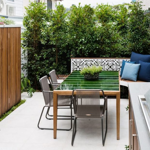 A compact landscaped outdoor courtyard dining area with a rectangular timber dining table topped by a glossy green surface, surrounded by modern woven metal-framed chairs. A built-in bench with blue cushions sits behind the table, paired with an ornate white patterned privacy screen. Tall dense hedging provides privacy, while a vertical timber slatted wall and pale tiled paving complete the space.