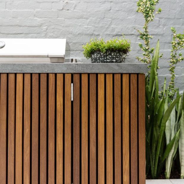 Close-up of a contemporary outdoor kitchen or built-in cabinet with vertical warm timber slats, a dark grey stone benchtop, and a stainless-steel appliance. The cabinet is set against a textured grey rendered brick wall with lush architectural planting alongside it.