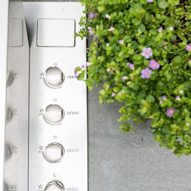 Close-up, top-down view of a stainless steel outdoor cooking appliance or built-in barbecue control panel, with multiple round control knobs and a rectangular display or cover. A flowering green plant partially overlaps the right side, and a grey textured surface surrounds the appliance.