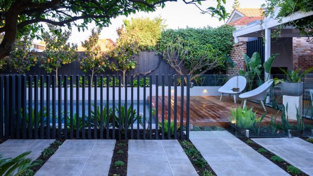A landscaped residential courtyard or front garden viewed from behind a dark vertical slat fence. Large pale concrete stepping slabs lead through planted garden beds toward a timber deck, with sculptural outdoor lounge chairs, dense hedging, mature trees, and a brick house with a tiled roof in the background.