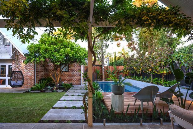 A landscaped residential courtyard viewed from beneath a covered pergola, featuring a timber deck, compact swimming pool, lush planting, lawn, stepping-stone path, brick walls, timber fencing, and outdoor seating. The garden is enclosed and private, with a strong indoor-outdoor connection.