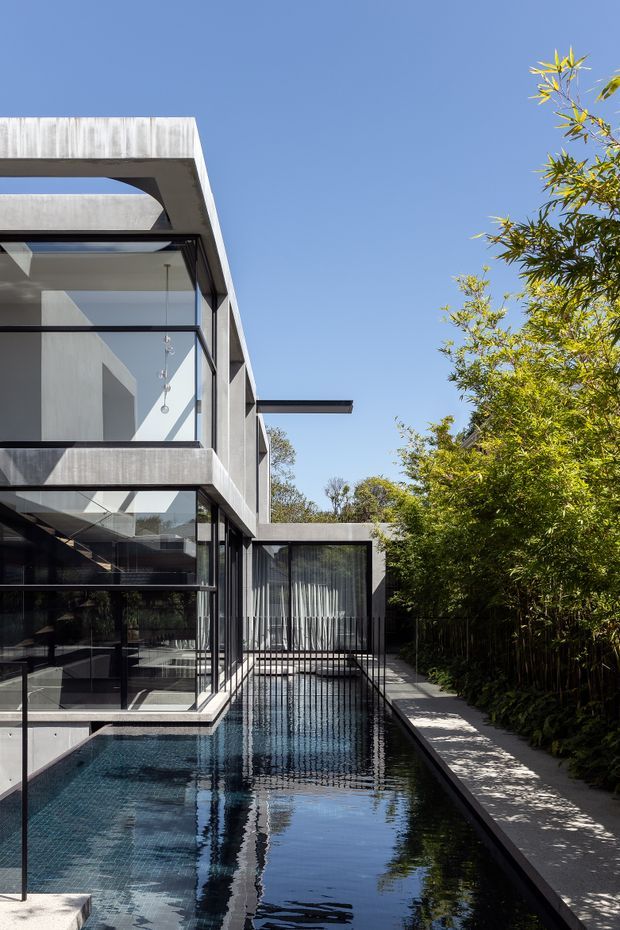 Exterior view of a contemporary multi-level residence with strong geometric concrete forms, dark metal-framed floor-to-ceiling glazing, and projecting flat roof elements. A long narrow reflective swimming pool runs alongside the house, bordered by a paved walkway and dense planted screening.