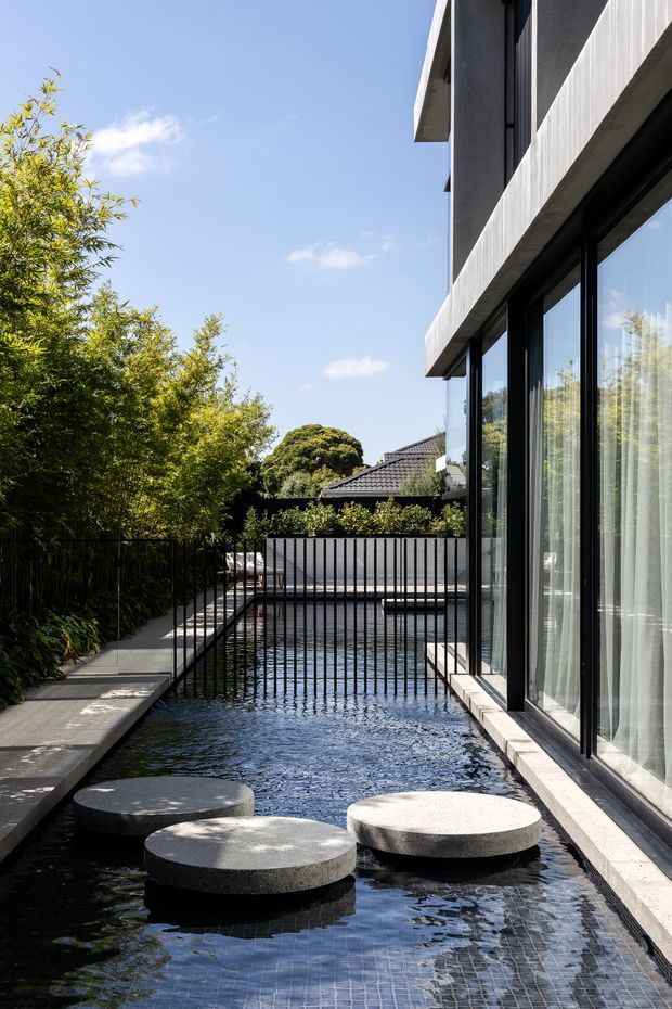 Contemporary residential exterior beside a narrow reflective water feature or pool, with large black-framed glazed openings, projecting concrete bands, and a paved walkway. Circular stepping stones cross the water, while mature planting and a screened boundary create a private courtyard-like setting.